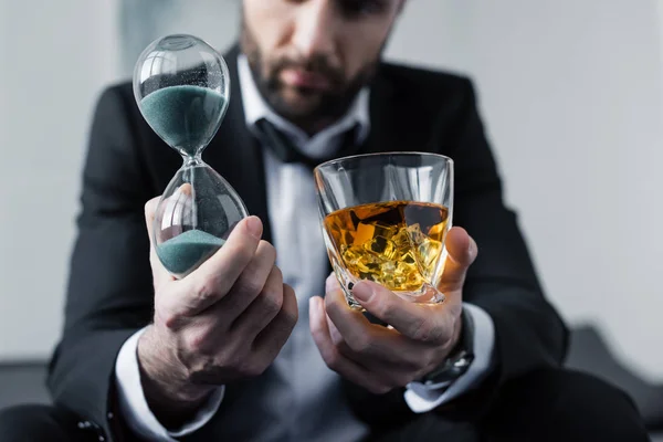 Vista recortada de empresario deprimido sosteniendo reloj de arena y vaso de whisky - foto de stock