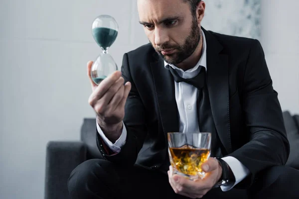 Enfoque selectivo de hombre de negocios molesto mirando reloj de arena mientras sostiene el vaso de whisky - foto de stock