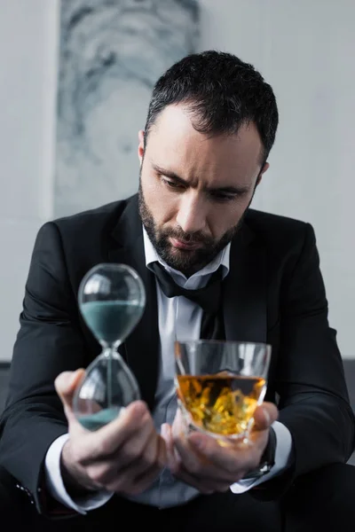 Избирательный фокус угнетенного бизнесмена, держащего стакан виски и песочные часы — стоковое фото