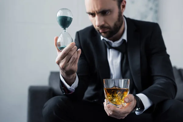 Selektiver Fokus eines aufgebrachten bärtigen Mannes im Anzug, der auf Sanduhr schaut, während er ein Glas Whiskey in der Hand hält — Stockfoto