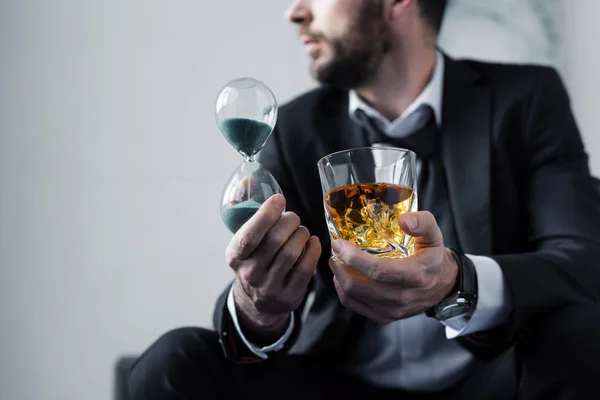 Partial view of adult man in suit holding hourglass and glass of whiskey — Stock Photo