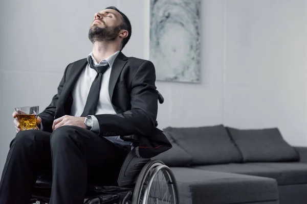 Bouleversé homme handicapé barbu assis en fauteuil roulant avec les yeux fermés et tenant un verre de whisky — Photo de stock