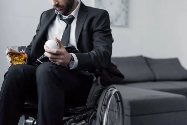 Vista recortada del hombre discapacitado en traje negro sentado en silla de ruedas con bola y vaso de whisky - foto de stock