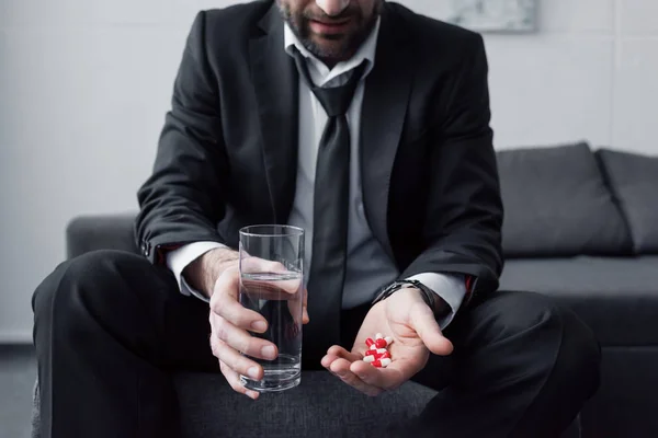 Cropped view of man in black suit holding glass of water and handful of pills — Stock Photo