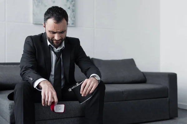 Depressed bearded man sitting on sofa while holding gift box and flask — Stock Photo