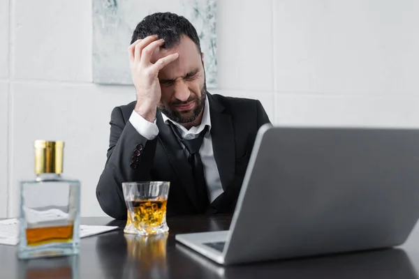 Molesto hombre de negocios sentado en el lugar de trabajo cerca de la computadora portátil, botella y vaso de whisky - foto de stock