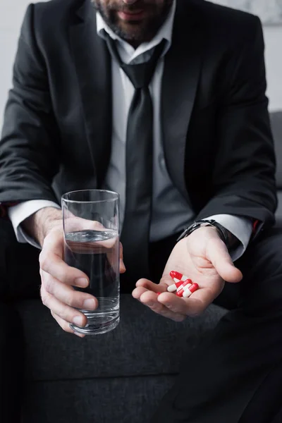 Partial view of depressed man holding glass of water and handful of pills — Stock Photo