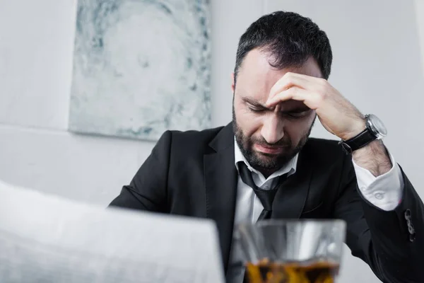 Hombre de negocios deprimido sosteniendo periódico mientras está sentado en el lugar de trabajo cerca de un vaso de whisky - foto de stock