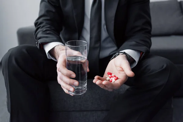 Vista recortada del hombre en traje negro sosteniendo un vaso de agua y un puñado de pastillas - foto de stock