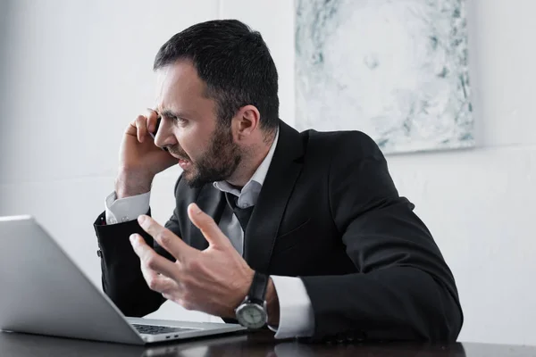 Homem de negócios zangado brigando enquanto sentado no local de trabalho e falando no smartphone — Fotografia de Stock