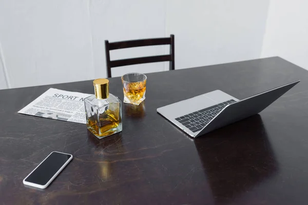 Scrivania in legno con computer portatile, giornale sportivo, smartphone con schermo bianco, bottiglia e bicchiere di whisky — Foto stock
