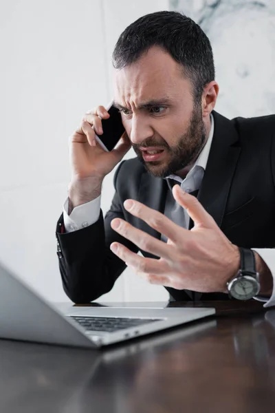 Hombre de negocios enojado peleando mientras habla en el teléfono inteligente cerca de la computadora portátil - foto de stock