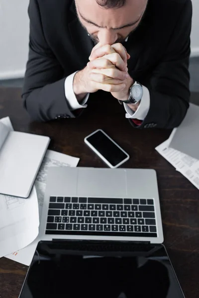 Vista de ángulo alto del hombre de negocios sentado en el lugar de trabajo cerca de la computadora portátil, papeles y teléfono inteligente con pantalla en blanco - foto de stock