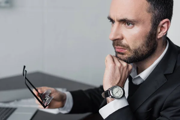 Upset bearded businessman sitting at workplace, holding glasses and looking away — Stock Photo