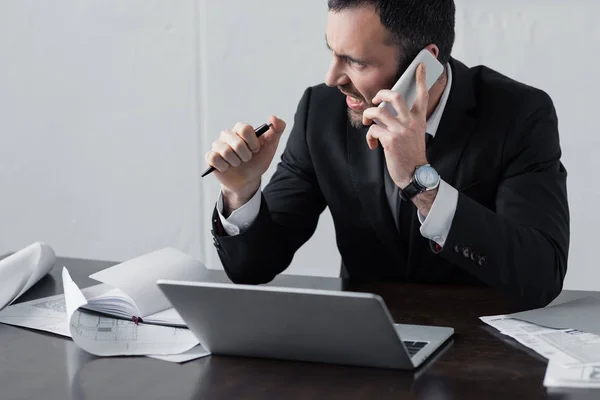 Unzufriedener Geschäftsmann im schwarzen Anzug spricht am Arbeitsplatz auf Smartphone — Stockfoto