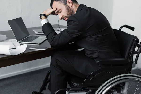 Hombre de negocios discapacitado deprimido en silla de ruedas sentado en el lugar de trabajo con computadora portátil, teléfono inteligente y papeles - foto de stock