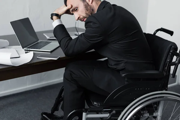 Molesto empresario discapacitado sentado en el lugar de trabajo en silla de ruedas y cogido de la mano en la frente - foto de stock