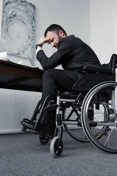 Vista de ángulo bajo del empresario discapacitado deprimido sentado en silla de ruedas en el lugar de trabajo con la cabeza inclinada - foto de stock