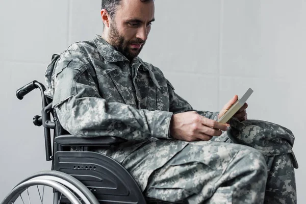 Nachdenklicher behinderter unrasierter Mann in Militäruniform sitzt im Rollstuhl und betrachtet Foto im Rahmen — Stockfoto
