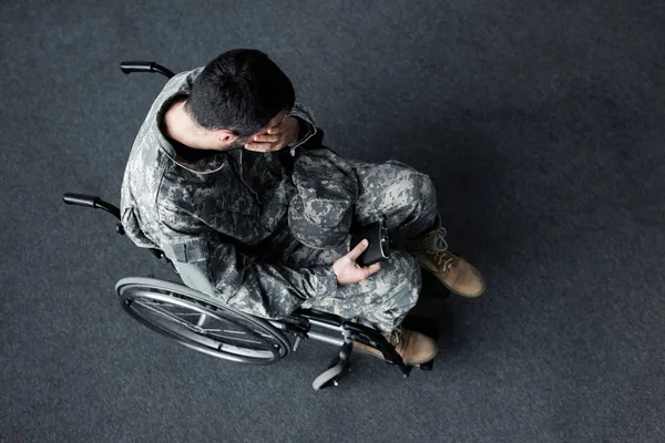 Vista aérea del hombre discapacitado en uniforme militar sentado en silla de ruedas y cubriendo la cara con la mano - foto de stock