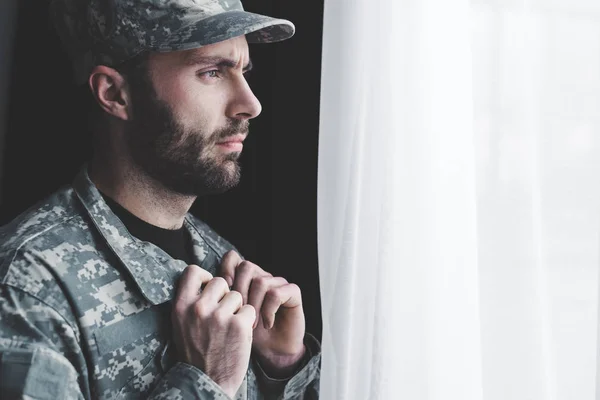 Hombre barbudo reflexivo en uniforme militar de pie junto a la ventana y mirando hacia otro lado - foto de stock