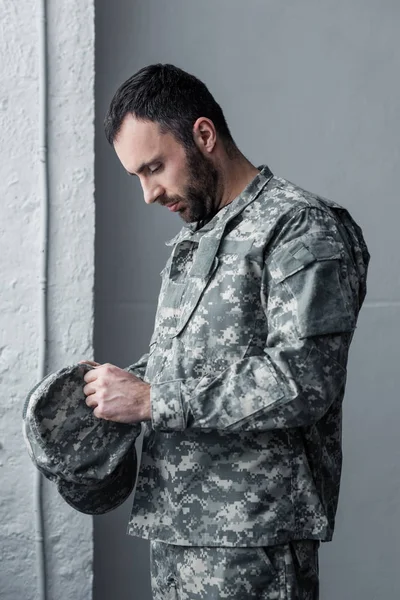 Triste barbudo hombre en uniforme militar de pie con arco tenía y la celebración de la tapa en las manos - foto de stock