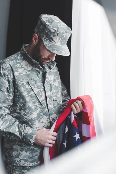 Foco seletivo de homem militar triste em uniforme segurando bandeira nacional dos EUA enquanto estava à janela com a cabeça curvada — Fotografia de Stock
