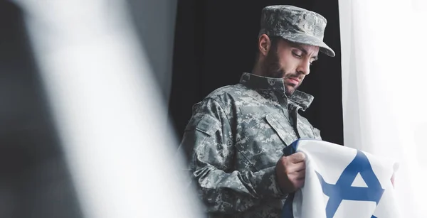 Selective focus of pensive military man in uniform holding israel national flag while standing by window — Stock Photo