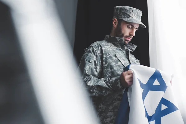 Enfoque selectivo del militar reflexivo en uniforme que sostiene la bandera nacional de Israel mientras está de pie por la ventana - foto de stock