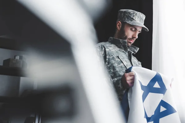 Messa a fuoco selettiva di uomo barbuto pensieroso in uniforme militare con bandiera nazionale israeliana mentre in piedi alla finestra — Foto stock