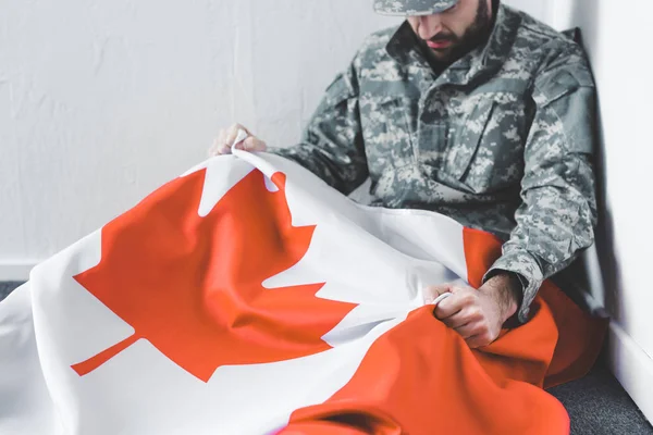 Hombre deprimido en uniforme militar sentado en el suelo en la esquina y sosteniendo la bandera nacional de Canadá - foto de stock