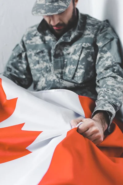 Enfoque selectivo del hombre deprimido en uniforme militar sentado en esquina y sosteniendo la bandera nacional de Canadá - foto de stock