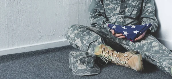 Обрезанный вид человека в военной форме, сидящего на полу в углу и держащего национальный флаг США — стоковое фото