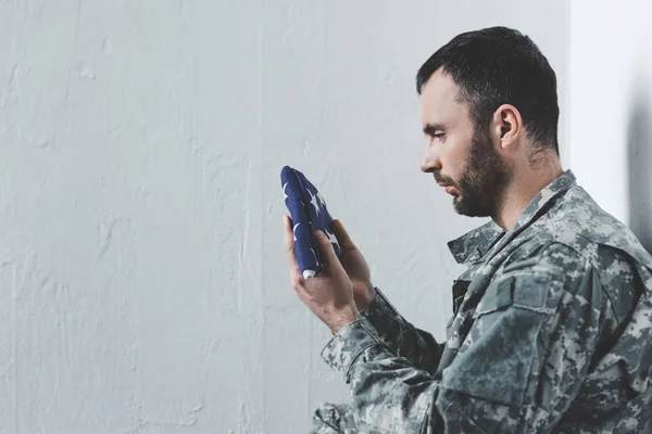 Vista laterale dell'uomo barbuto in uniforme militare seduto vicino al muro bianco e con bandiera nazionale degli Stati Uniti — Foto stock