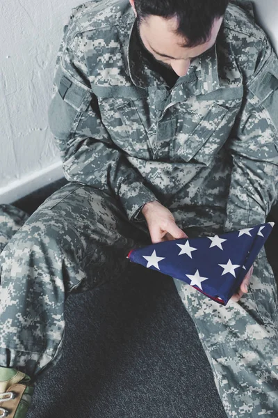 High angle view of depressed man in military uniform sitting on floor in corner and holding usa national flag — Stock Photo