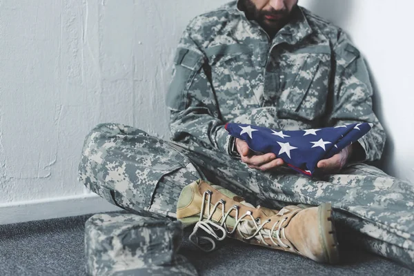 Vista parcial del hombre deprimido en uniforme militar sentado en el piso gris en la esquina y sosteniendo la bandera nacional de EE.UU. - foto de stock