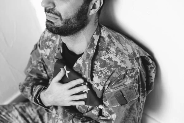 Обрезанный вид депрессивного военного человека, сидящего у белой стены и держащего США военный человек рядом с сердцем — стоковое фото