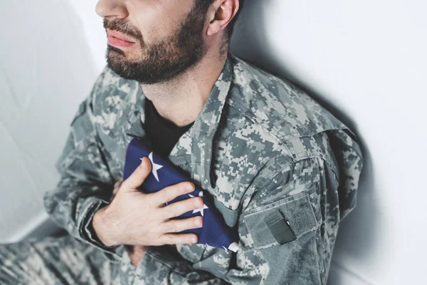 Partial view of depressed military man sitting by white wall and holding usa military man near heart — Stock Photo