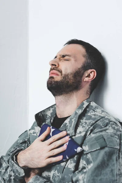 Depressiver Soldat sitzt mit geschlossenen Augen an weißer Wand und hält US-Nationalflagge in der Hand — Stockfoto