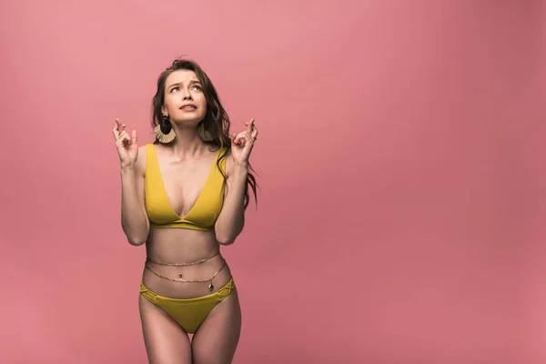 Chica preocupada en traje de baño amarillo mirando hacia arriba con los dedos cruzados aislados en rosa - foto de stock