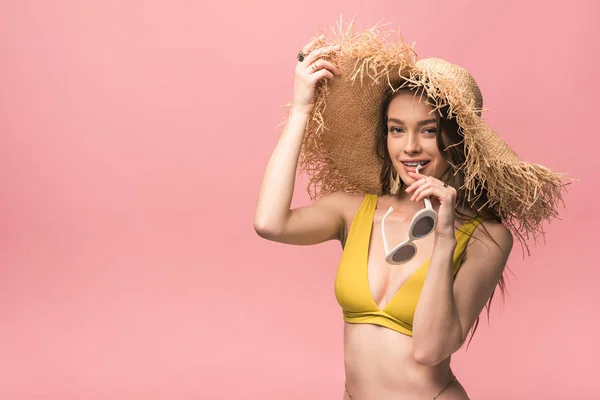 Smiling girl in yellow swimsuit and straw hat holding sunglasses isolated on pink — Stock Photo