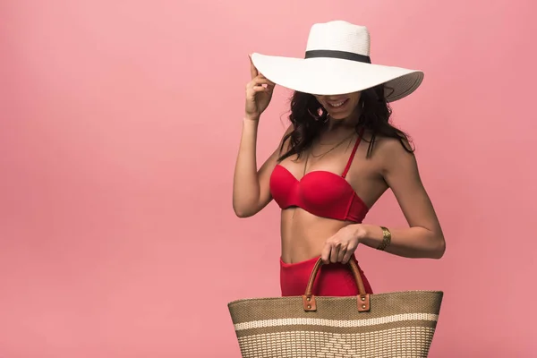Усміхнена приваблива жінка в купальнику і капелюсі тримає пляжний мішок ізольовано на рожевому — стокове фото