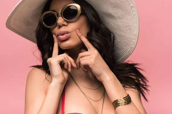 Attraktive junge Frau mit Hut und Sonnenbrille berühren Gesicht isoliert auf rosa — Stockfoto