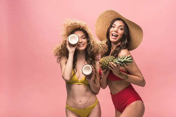 Две улыбающиеся девушки в соломенных шляпах держат кокосы и ананас изолированными на розовом — стоковое фото