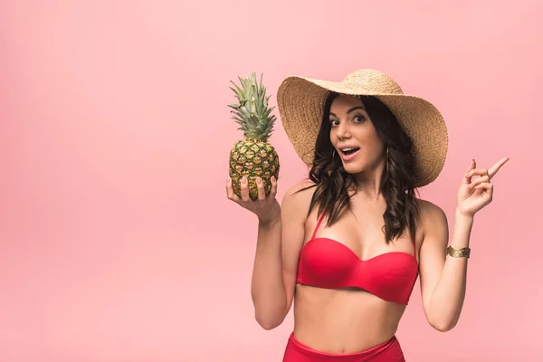 Amazed girl in straw hat and bikini holding pinapple isolated on pink — Stock Photo