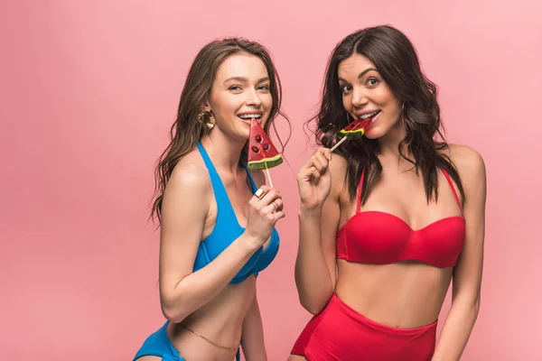 Dos chicas guapas en bikini comiendo piruletas de sandía aisladas en rosa - foto de stock