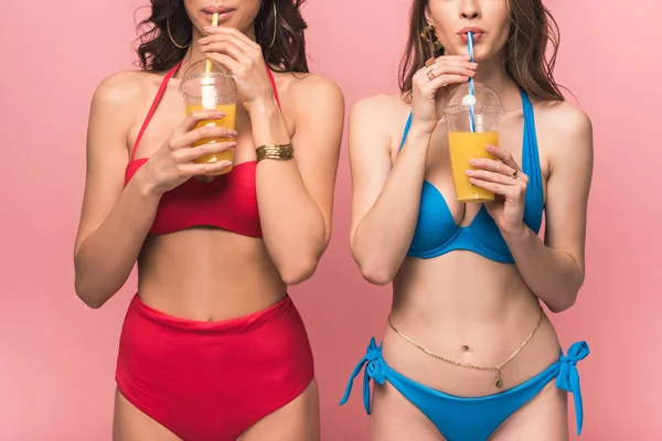 Vista ritagliata di due belle ragazze in bikini che bevono succo d'arancia isolato su rosa — Foto stock