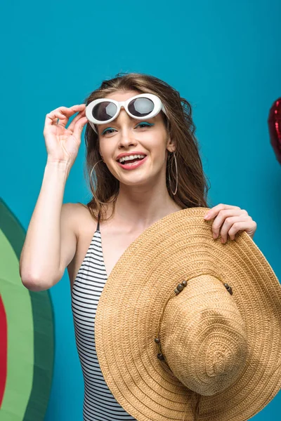 Heureuse jeune femme en lunettes de soleil tenant chapeau de paille sur bleu — Photo de stock