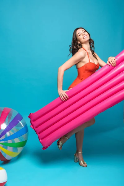 Full length view of smiling woman in swimsuit holding inflatable swimming mattress on blue — Stock Photo