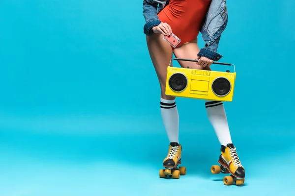 Cropped view of woman in swimsuit and roller skates holding boombox and cassette tape on blue — Stock Photo